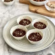 chocolate hazelnut tartlets served on a round plate, garnished with freshly chopped hazelnuts, with a bowl of hazelnuts and glass of milk in the background.
