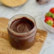 healthy chocolate tahini spread served in jar on wooden board, with sesame seeds scattered around the board.