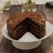 rich chocolate gateau with a few slices missing, served on white cake stand, glasses of milk in the background.