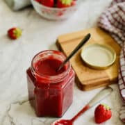 homemade strawberry jam served in a jar with a tempting spoonful of jam, fresh strawberries in the background.