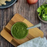 birds eye view of fresh green coriander chutney served in a bowl on a wooden board, coriander leaves, red apples and ginger in the background.