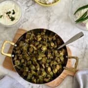 birds eye view of stir-fried okra (bhindi ki sabzi) on board, with rice in background, raita and onions and chillies in background.