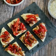 Mediterranean flatbread with naan, hummus and roasted onions and peppers served on black board, with ingredients in background.