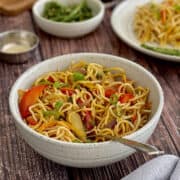 fresh and delicious vegetable hakka noodles in a bowl, with a fork, plate of food in background.