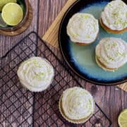 overhead view of lime and coconut layered cupcakes on wire rack and blue plate, with fresh limes in background.