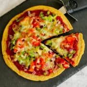 fresh vegetable chickpea flour pizza partly sliced and served on black serving plate, with a pizza cutter.