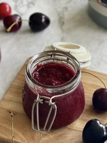 fresh homemade cherry jam, served in a jar, with cherries in the background.