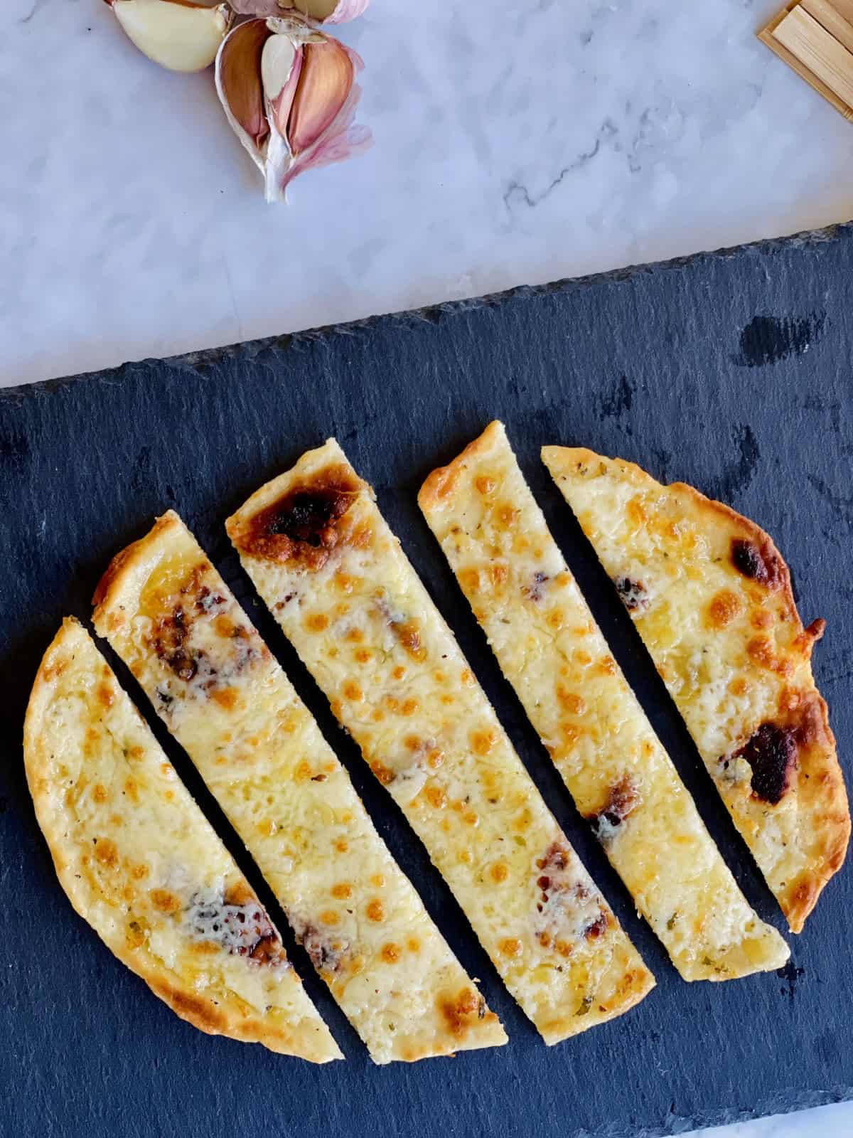 freshly made golden cheesy garlic naan bread slices served on a black plate, with fresh garlic in the background.