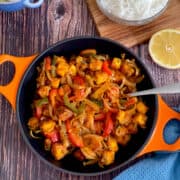 chilli paneer in a serving pan, with lemon and rice and a blue cloth in the background.