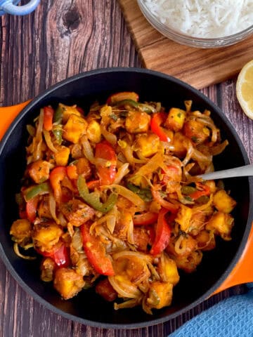 chilli paneer in a serving pan, with lemon and rice and a blue cloth in the background.