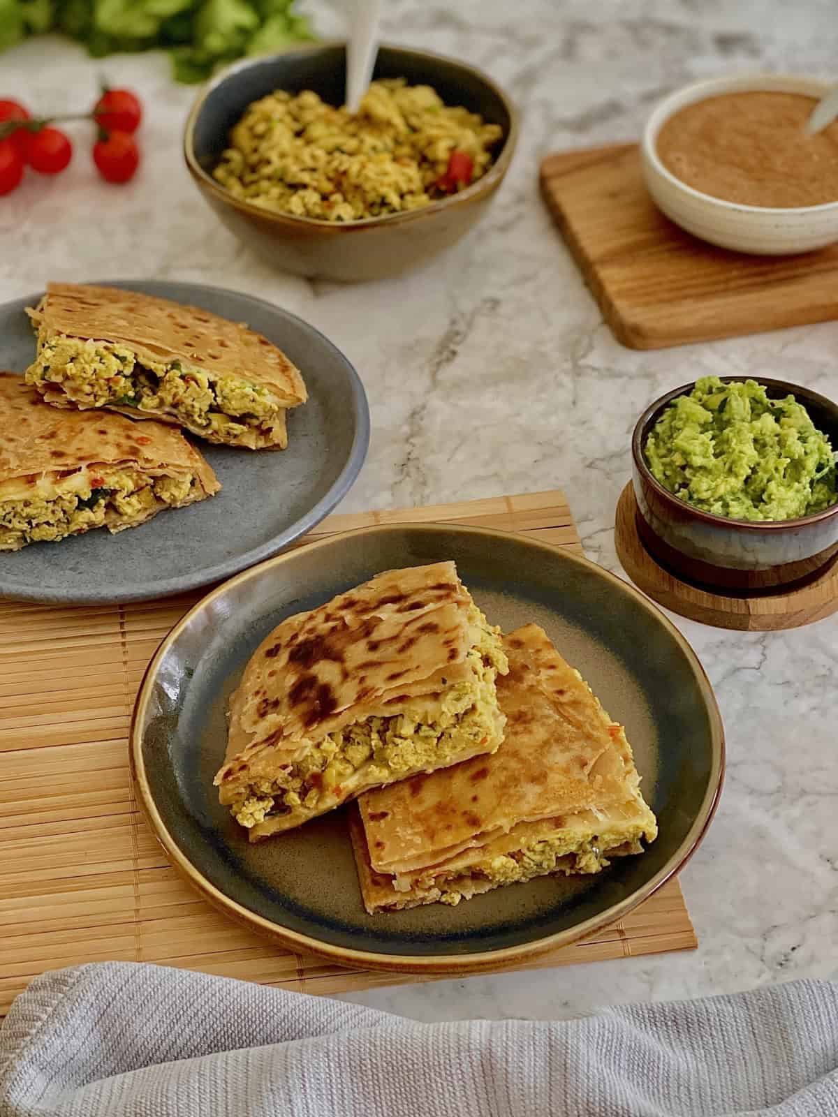 crispy masala egg paratha quarters on a plate, with fresh guacamola, salsa, tomatoes on the vine, in the background.