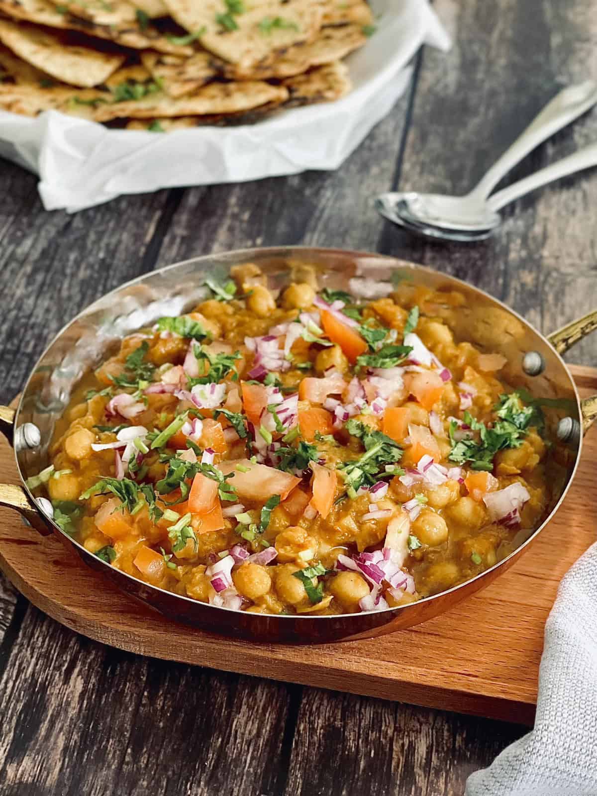 Chickpea masala served in a serving dish, garnished with fresh coriander, tomatoes, onions, a basket of naan in the background.