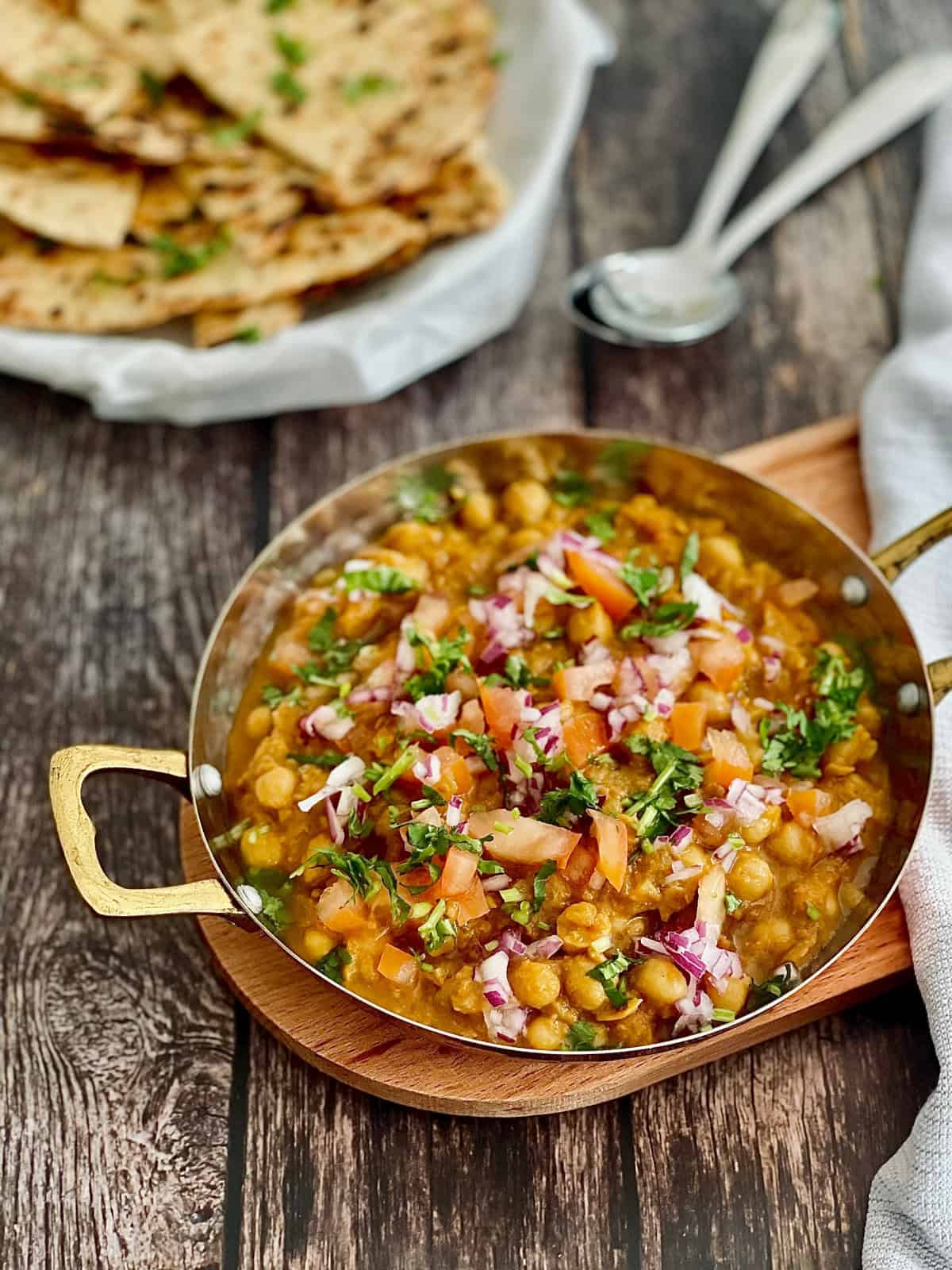 Chickpea masala served in a serving dish, garnished with fresh coriander, tomatoes, onions, a basket of naan and spoons in the background.