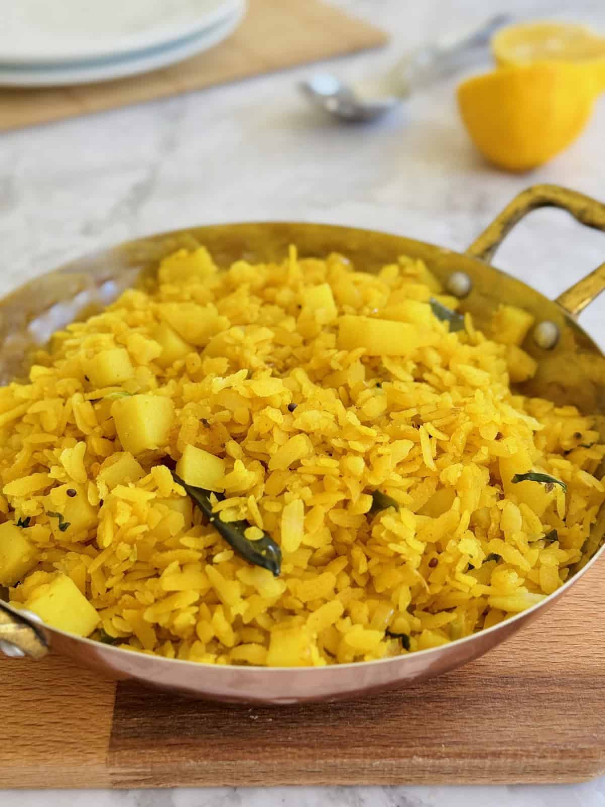 fresh bright yellow poha in serving dish, with lemon wedges and spoons nearby.